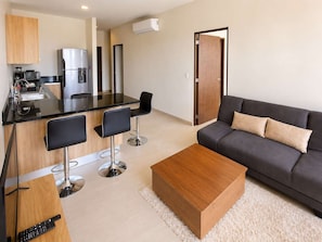 A modern living room with a sofa, a coffee table, and a kitchen area with a refrigerator, microwave, and sink.