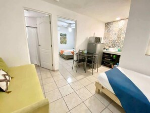 A compact hotel room with a bed, a desk, a chair, a small kitchenette, and a bathroom.