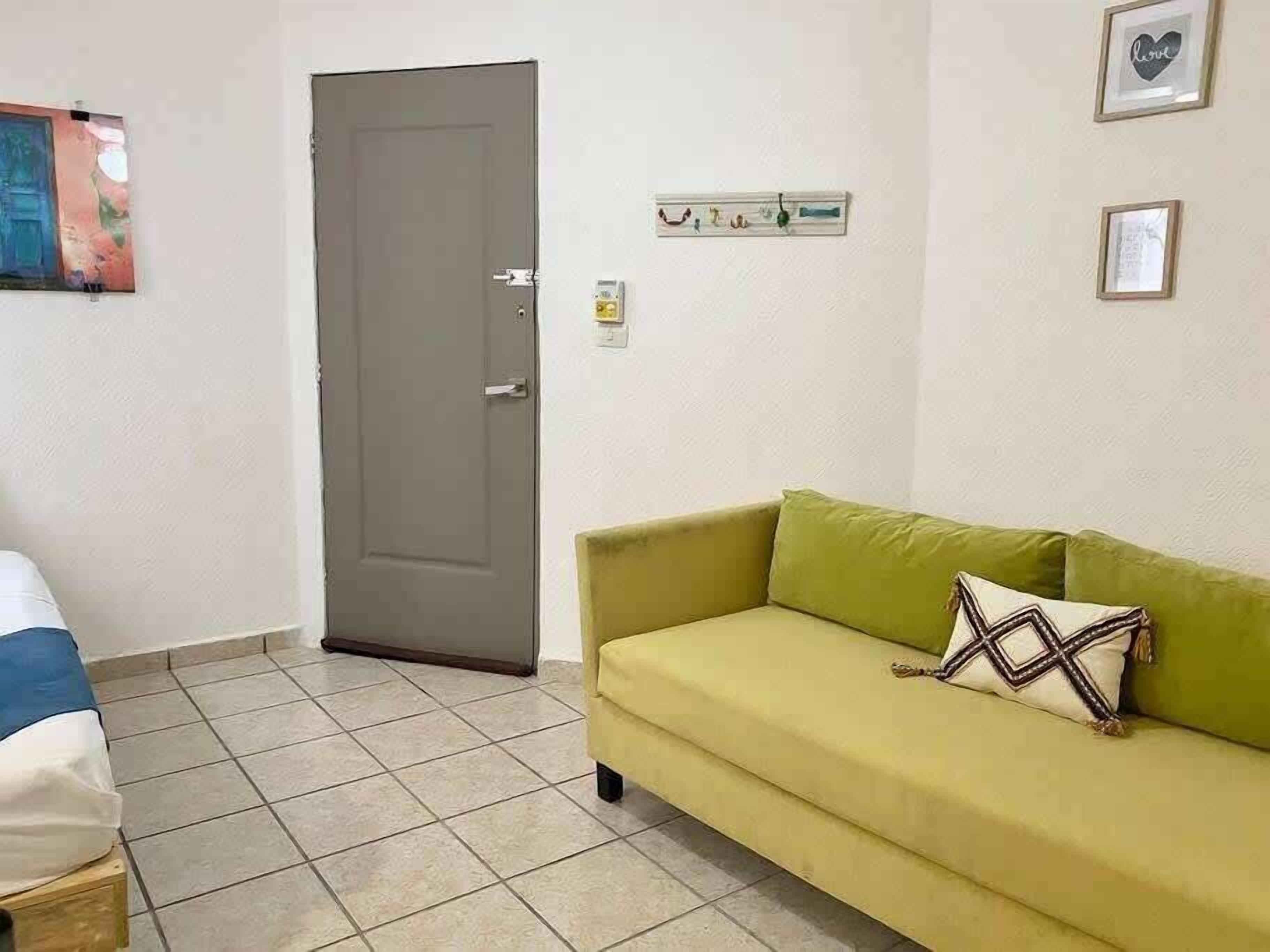Quarto com porta cinzenta, sofá amarelo com almofada estampada, cama com cobertura azul e branca, e prateleira fixada na parede com objetos pequenos.