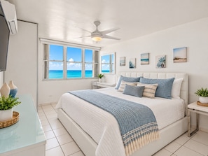 A bedroom with a large bed, a ceiling fan, a desk, and a view of the ocean.