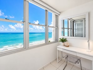 Una habitación con un ventanal grande con vista a una playa, un escritorio blanco y una silla.
