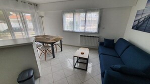 Ein Wohnzimmer mit blauem Sofa, kleinem Tisch, Esstisch mit Stühlen und Fenster mit Blick auf Gebäude.