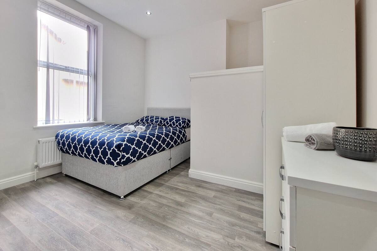 A bedroom with a bed, a window, a radiator, a wardrobe, and a small table with a bowl.