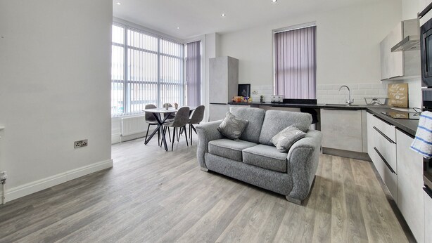 A modern kitchen with white cabinets, a black countertop, and a built-in oven. There is a dining area with a round table and chairs, and a grey sofa in the living room. - Dysa Waterloo Apartments (Blackpool)