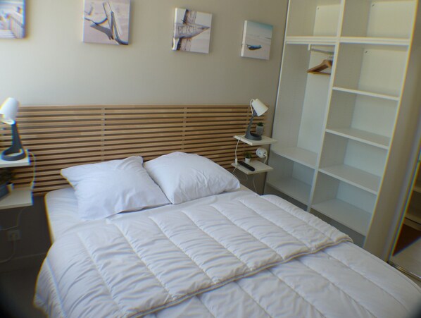 Una cama bien hecha con ropa de cama blanca, una mesita de noche con una lámpara y otra mesita con un mando a distancia y una planta pequeña.