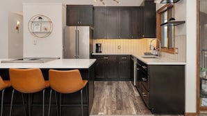 A modern kitchen with dark cabinets, a stainless steel refrigerator, a coffee maker, and a bar area with stools. - Amazing Views! Downtown Penthouse with Sauna (Winter Park)