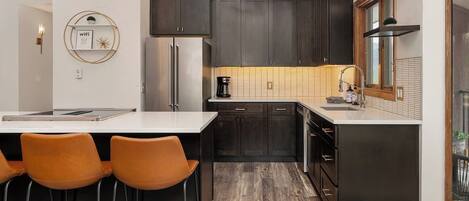 A modern kitchen with dark cabinets, a stainless steel refrigerator, a coffee maker, and a bar area with stools.