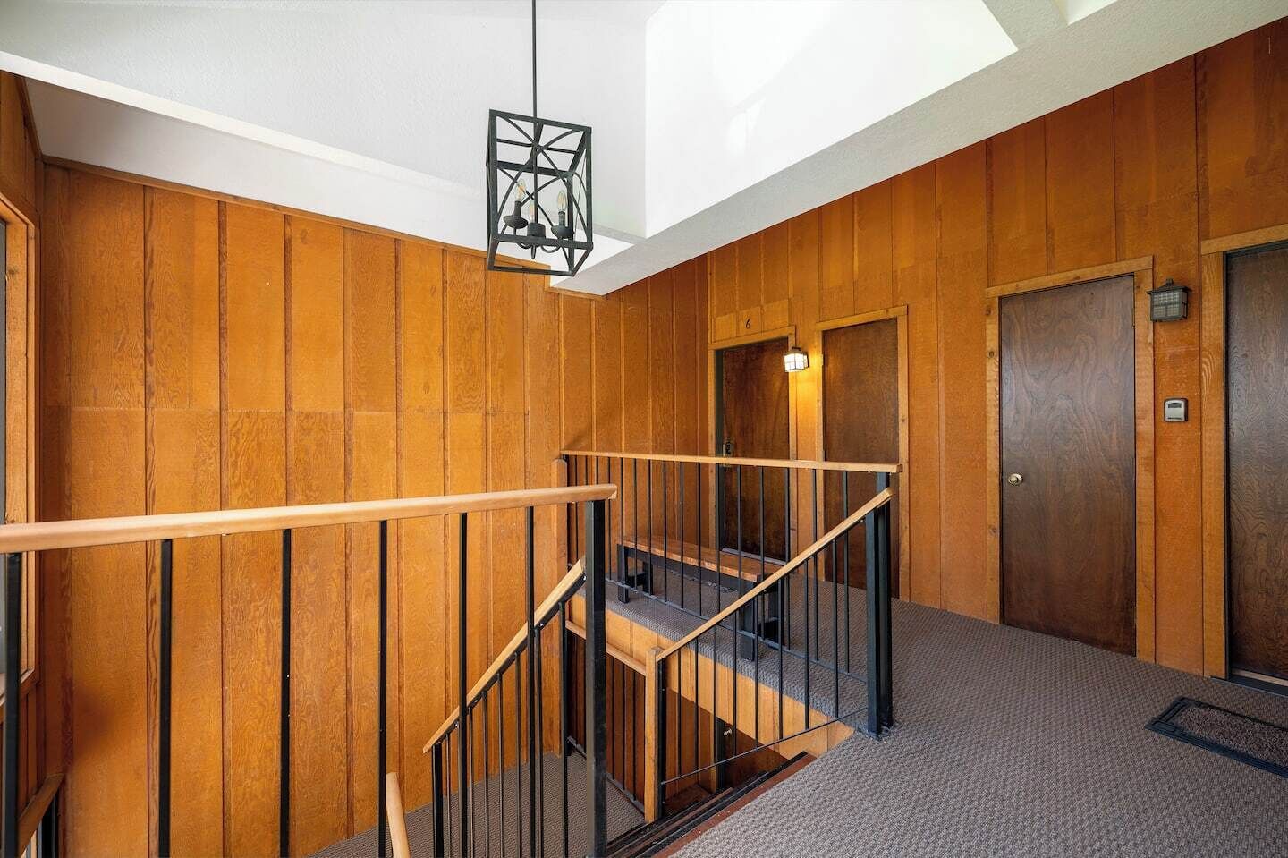 Un couloir aux murs lambrés, avec un escalier, un luminaire suspendu et une porte dotée d’un interrupteur.