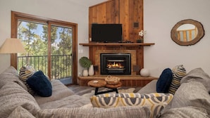 A cozy living room with a fireplace, a television, and a balcony with a view of trees. - Amazing Views! Downtown Penthouse with Sauna (Winter Park)