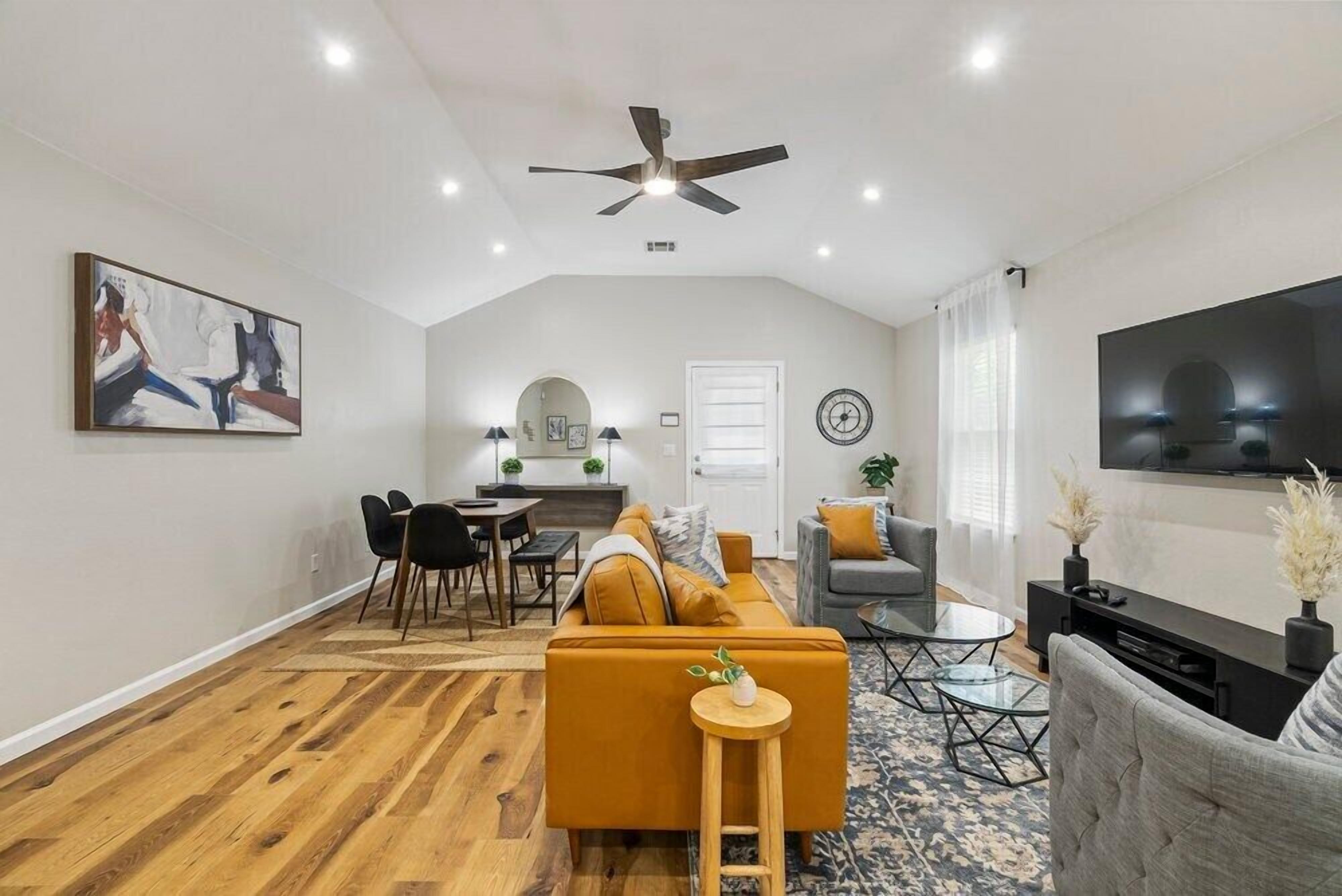 Een moderne woonkamer met een bank, salontafel en televisie. Er is een eethoek met stoelen en een tafel. De kamer heeft houten vloeren en een plafondventilator.