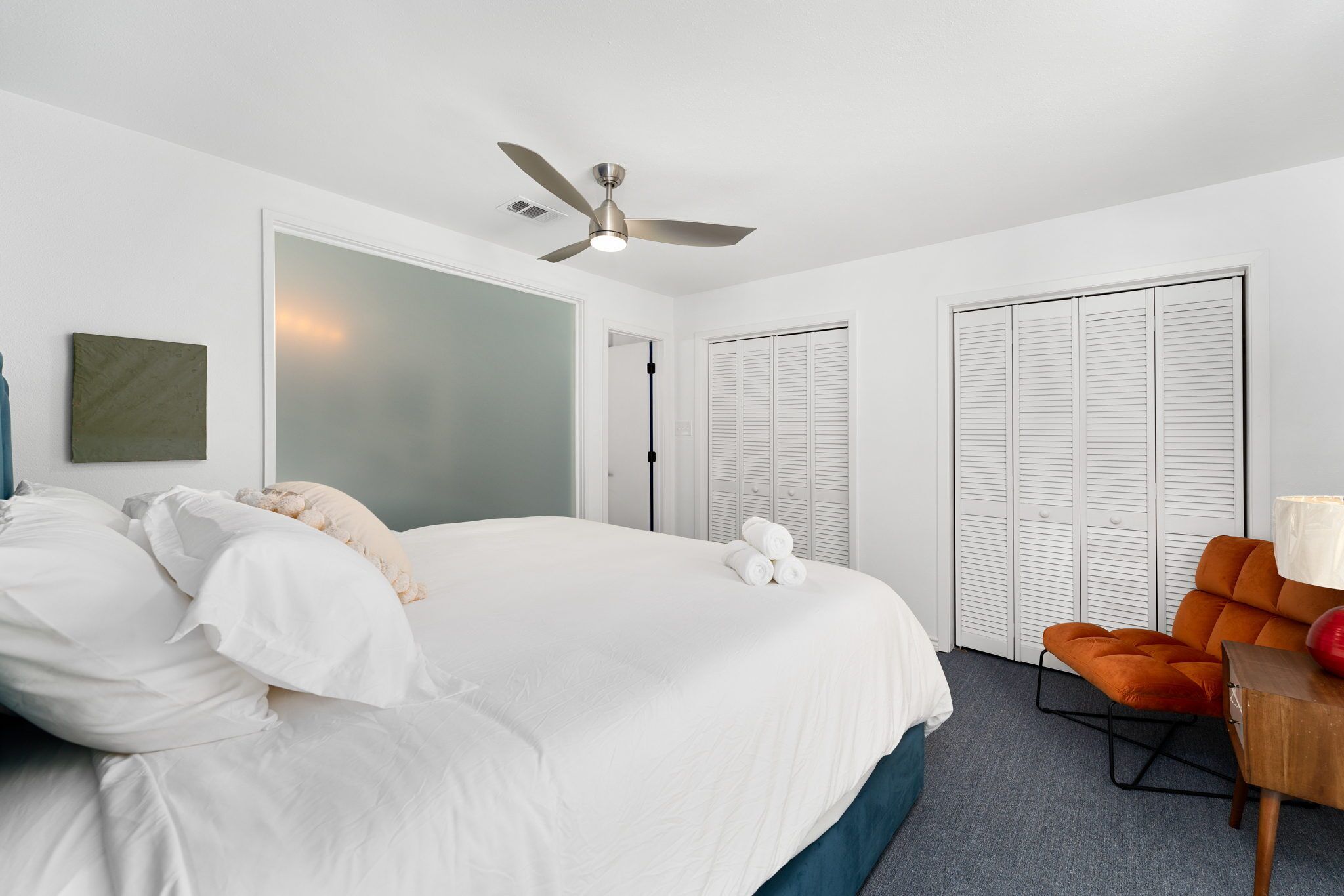 A bedroom with a large bed, a ceiling fan, white closets, and a small orange chair.