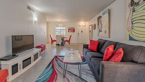 Ein modernes Wohnzimmer mit einem Flachbildfernseher, einem grauen Sofa mit roten Kissen, einem Couchtisch und einem farbenfrohen Teppich.