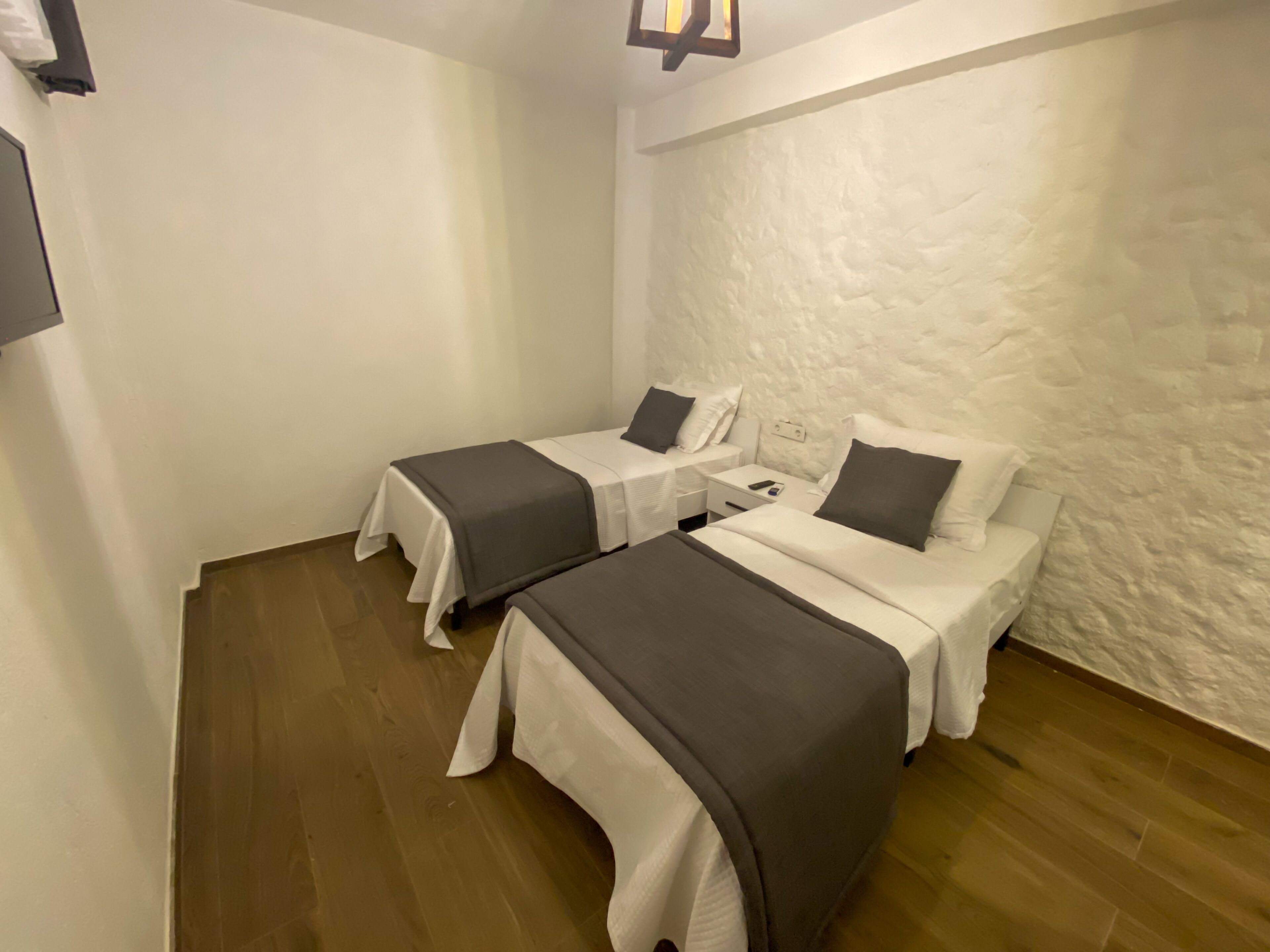 Ein Zimmer mit zwei Betten, jedes mit einer weißen Matratze und grauer Bettwäsche. Ein Holzboden und eine strukturierte weiße Wand.