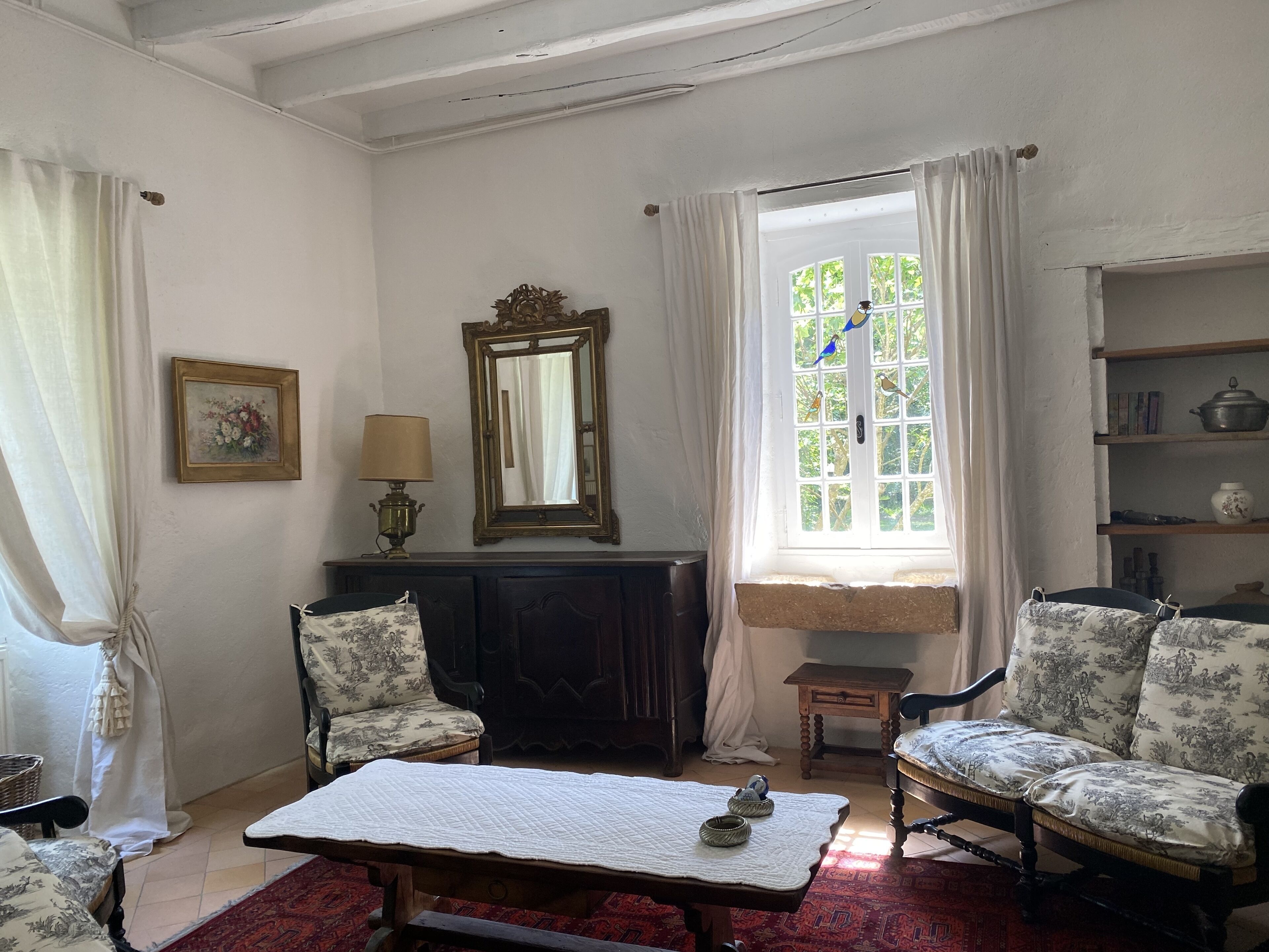 Una habitación tradicional con chimenea, dos sillones, una mesa y un ventanal con cortinas.