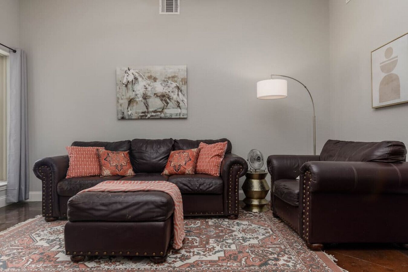 Ein Wohnzimmer mit einem dunkelbraunen Ledersofa, einem passenden Hocker und einem gemusterten Teppich. Außerdem steht eine Stehlampe im Raum und es hängt ein Gemälde an der Wand.