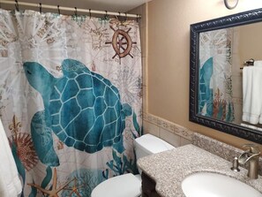 Un baño con cortina de ducha de tortuga marina, un adorno de timón de barco, encimera de granito y un espejo grande.