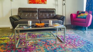 Een woonkamer met een bruine leren bank, een glazen salontafel met tijdschriften, een kleurrijk vloerkleed, een levendig schilderij aan de muur en een roze fauteuil.