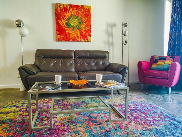 Una sala de estar con un sofá de cuero marrón, una mesa de centro de vidrio con revistas, una alfombra colorida, un cuadro llamativo en la pared y un sillón rosa.