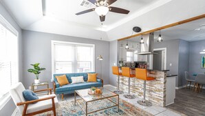 A modern living room with a blue sofa, a coffee table, and a kitchen area with a bar and stools. - Amazing Bungalow - by State Fair Grounds, WiFi, KG BD (Oklahoma City)