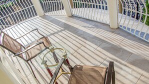 Uma área de estar coberta ao ar livre, com mesa de vidro e cadeiras de vime, delimitada por uma cerca de metal branco.