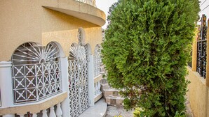 Um edifício com fachada curva, portões metálicos decorativos e um jardim bem cuidado com uma árvore alta e plantas em vasos.