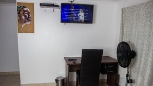 Une pièce comprenant un bureau, une chaise, un téléviseur et un ventilateur sur pied.