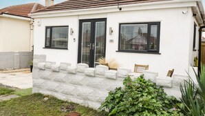 Einbungalow mit weißer Fassade, einem Ziegeldach und einem kleinen Vorgarten mit einer Bank und Kübelpflanzen.