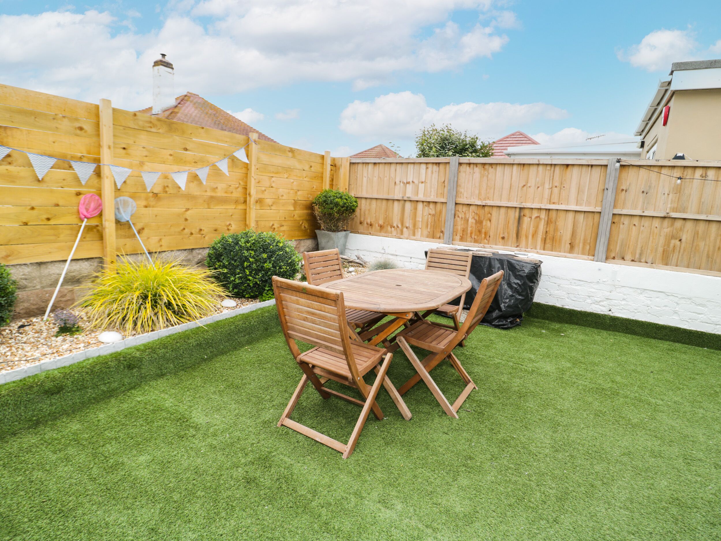 Patio con mesa y sillas de madera, rodeado de césped artificial y una valla de madera.