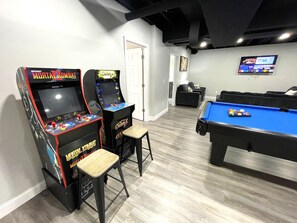 Ein Billardtisch, zwei Spielautomaten und ein Fernseher in einem Raum mit Holzboden und einer Tür im Hintergrund.