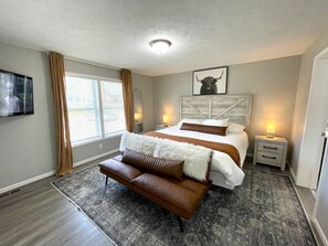 Ein Schlafzimmer mit einem großen Bett, einem Sofa, einem Nachttisch, einem Fernseher und einem Fenster mit Vorhängen.