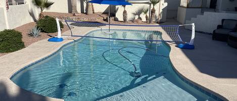 A swimming pool with a blue umbrella, surrounded by a landscaped area with palm trees and a house in the background.