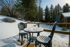 Un área de estar al aire libre cubierta de nieve con una mesa y sillas, rodeada por una valla de madera y una cabaña al fondo.