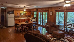 A rustic cabin interior with a wooden dining area, a sofa, and a large window overlooking a deck.