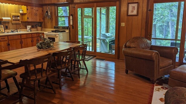 A wooden interior with a dining table, chairs, a sofa, and a grill outside the door.