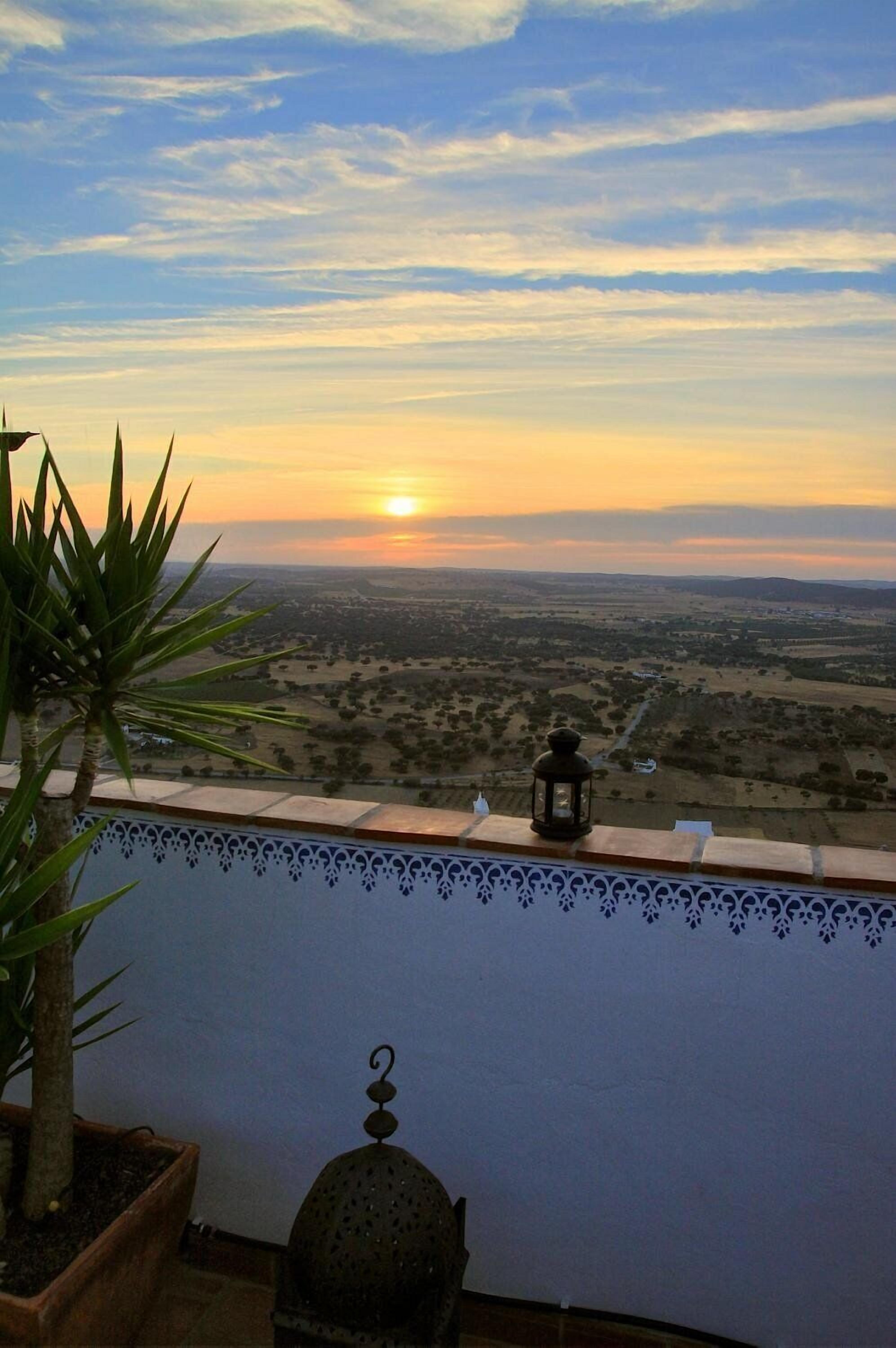 A balcony with a view of a sunset over a landscape, a potted plant, and a lantern.