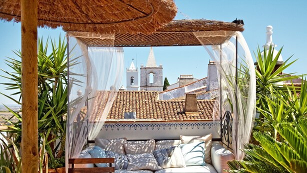 A cozy outdoor seating area with a thatched roof, cushioned seating, and a view of a church tower. - Casa pinto (Reguengos de Monsaraz)