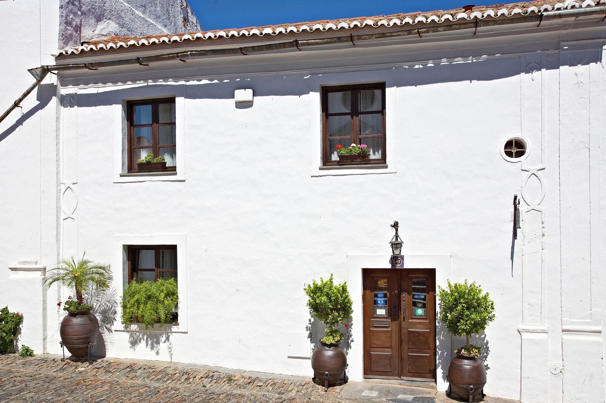 Un bâtiment blanc avec une porte en bois, deux fenêtres ornées de jardinières et une petite fenêtre ronde.