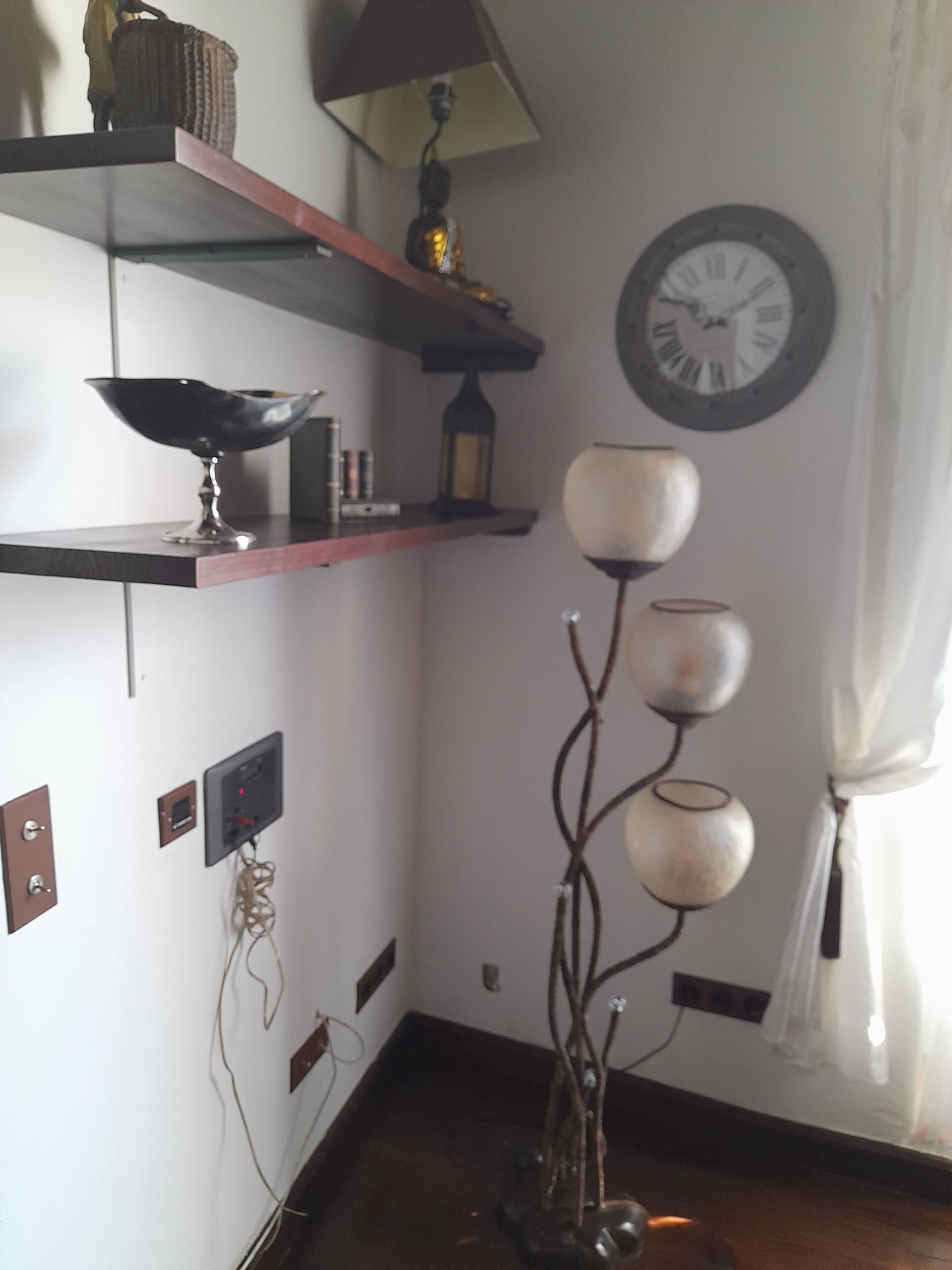 Een kamer met een houten vloer, een wandklok, planken met diverse spullen en een lamp.