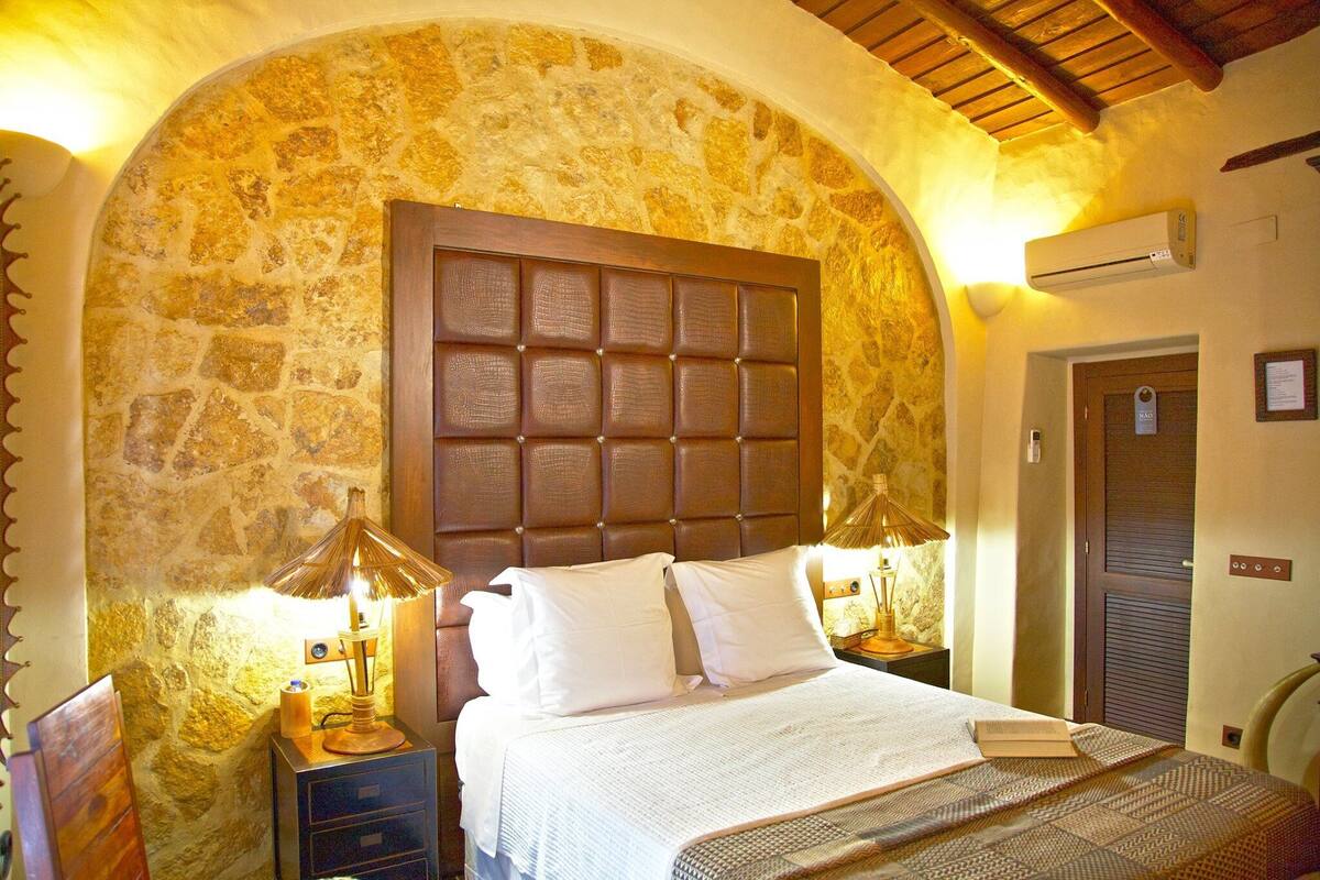 A bedroom with a stone wall, a wooden headboard, two bedside tables with lamps, and a door with a sign.