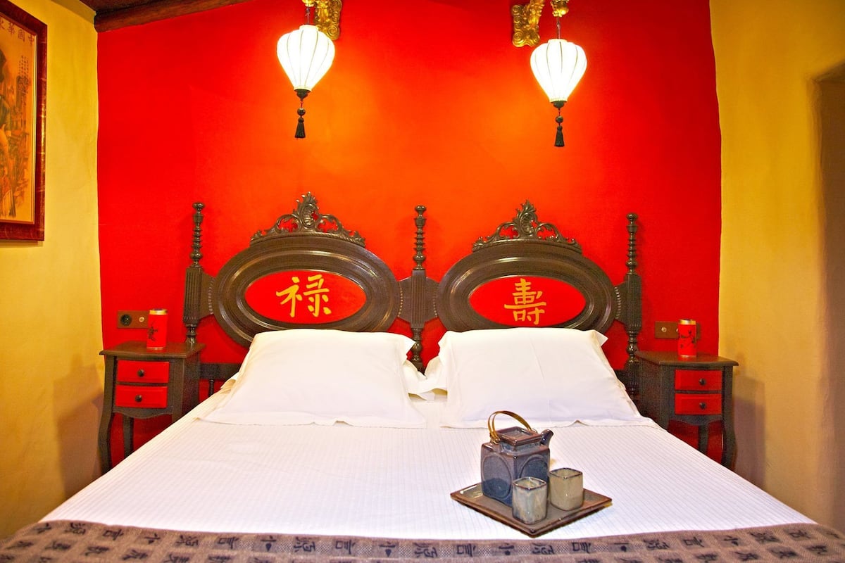 A hotel room with a red wall, a wooden bed frame, two bedside tables, and a nightstand with a lamp.