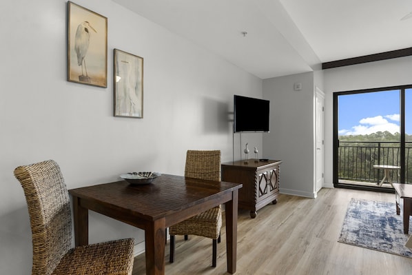 A dining area with a wooden table and wicker chairs, a flat-screen TV mounted on the wall, and a view of the outdoors through a large sliding glass door.