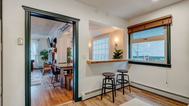 A modern interior with a wooden floor, a dining area with a table and chairs, a bar area with stools, and a large window with a view.