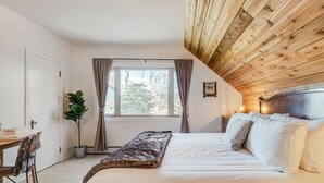A bedroom with a wooden ceiling, a large window with curtains, a bed with white linens, a bedside table, and a chair.