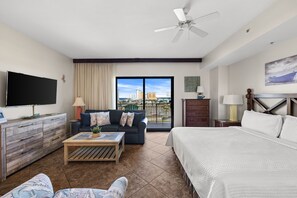 Habitación de hotel con cama, sofá, mesa de centro, televisor, ventilador de techo, balcón con vista a la ciudad y cómoda de madera.