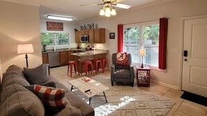 A living room with a sofa, a coffee table, a red chair, and a kitchen area with wooden cabinets and a ceiling fan. - Walk to Stadium/Strip/Downtown! 2 bd/2 bath, Off-street parking! (Tuscaloosa)