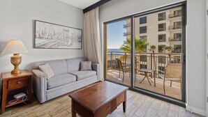 Ein modernes Wohnzimmer mit einem grauen Sofa, einem Holztisch, einer Lampe und einem großen Fenster mit Blick auf einen Balkon und Gartenmöbel.
