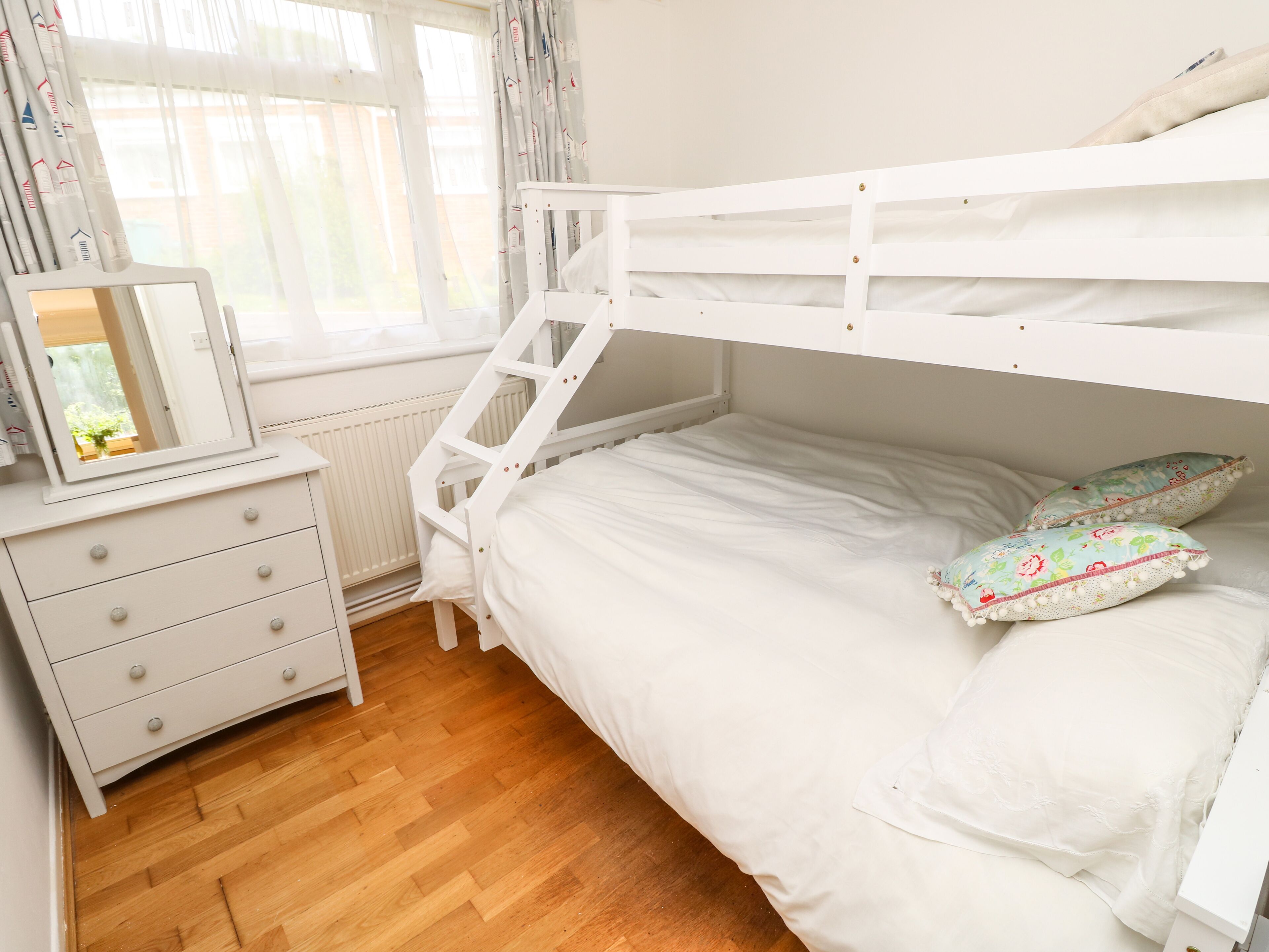 A bunk bed with white bedding, a dresser with drawers, a mirror, and two decorative pillows.