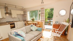 Una sala de estar acogedora con un sofá, una mesa de madera y una silla. Hay una zona de cocina con estufa y armarios. Se ve una ventana con cortinas, y una puerta da a un área exterior.