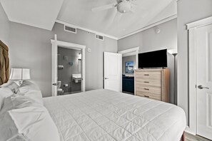 Ein Schlafzimmer mit einem großen Bett, einem Deckenventilator, einem Schminktisch, einem Fernseher und einer Tür zu einem Badezimmer.