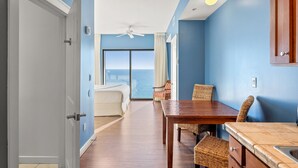 A hotel room with a bed, a dining table, wicker chairs, a view of the ocean, and a ceiling fan.